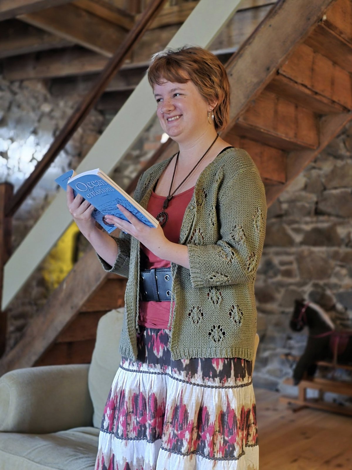 Lily O. Laverick at a Creative Slam A smiling woman called Lily O. Laverick, stands in a cozy room holding a blue book, written by her. She wears a green knit cardigan and a floral skirt, with a wooden staircase behind her.